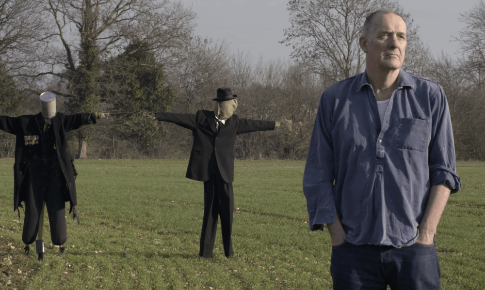 Bill Drummond stands with two scarecrows