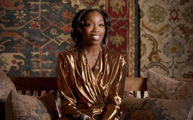 Estelle (present time) being interviewed while dressed in a shiny bronze-colored outfit, in front of rustic background with carpets on the walls.