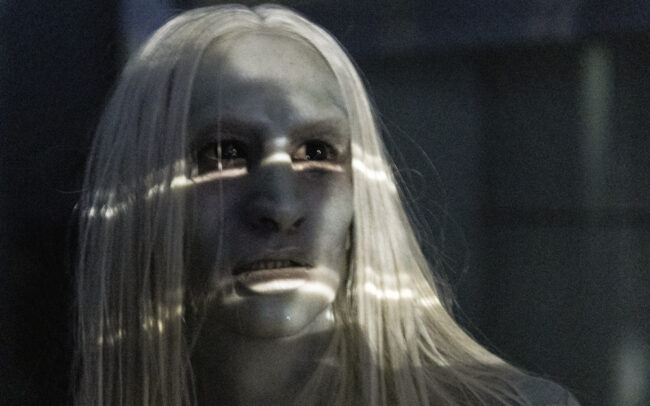 Close-up still from All You Need Is Death with actor Ben Stewardson looking off camera towards a source of light beaming white light across his face, with long white hair and dark black eyes.