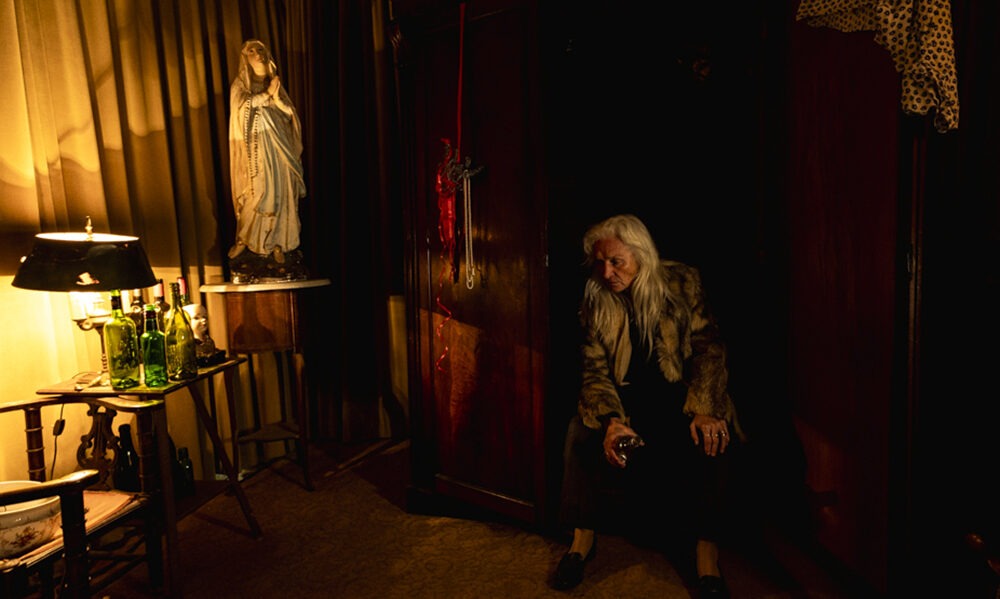 Still from All You Need Is Death where actress Olwen Fouere playing the singer who sings a cursed song is sat in a room with a large wooden wardrobe, bottles and a statue of the virgin Mary.